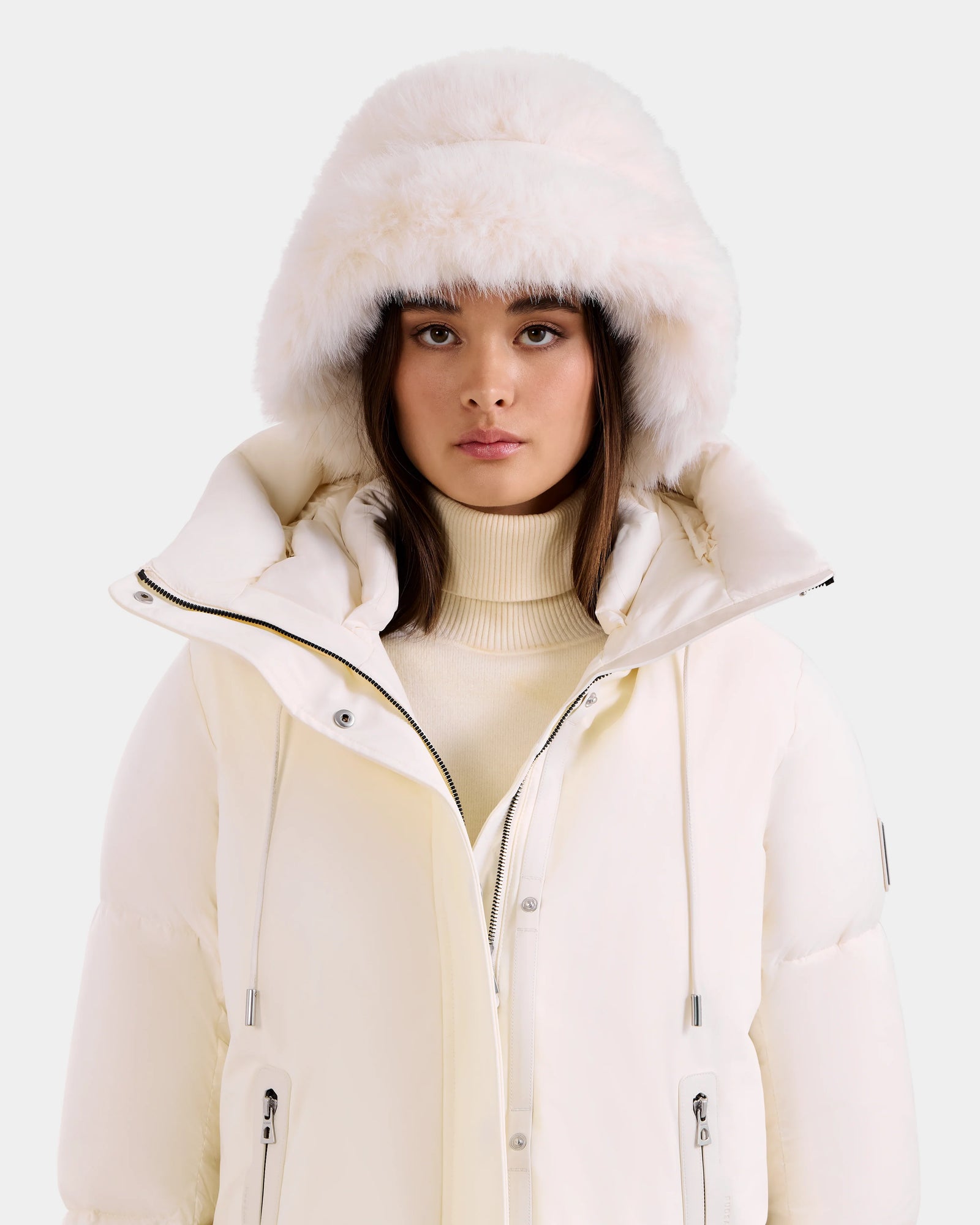 A woman models Rudsak’s YURI - L425538 Iceland white puffer jacket with vegan-leather trim, styled with a cream turtleneck sweater and faux-fur bucket hat—showcasing chic winter accessories against a plain light background.