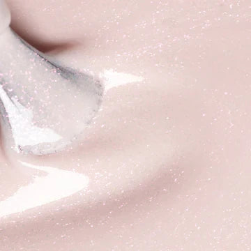 Close-up of a brush applying a shimmery, pale pink nail polish. The polish has a glossy, sparkling texture and the brush is partially visible in the top left corner.
