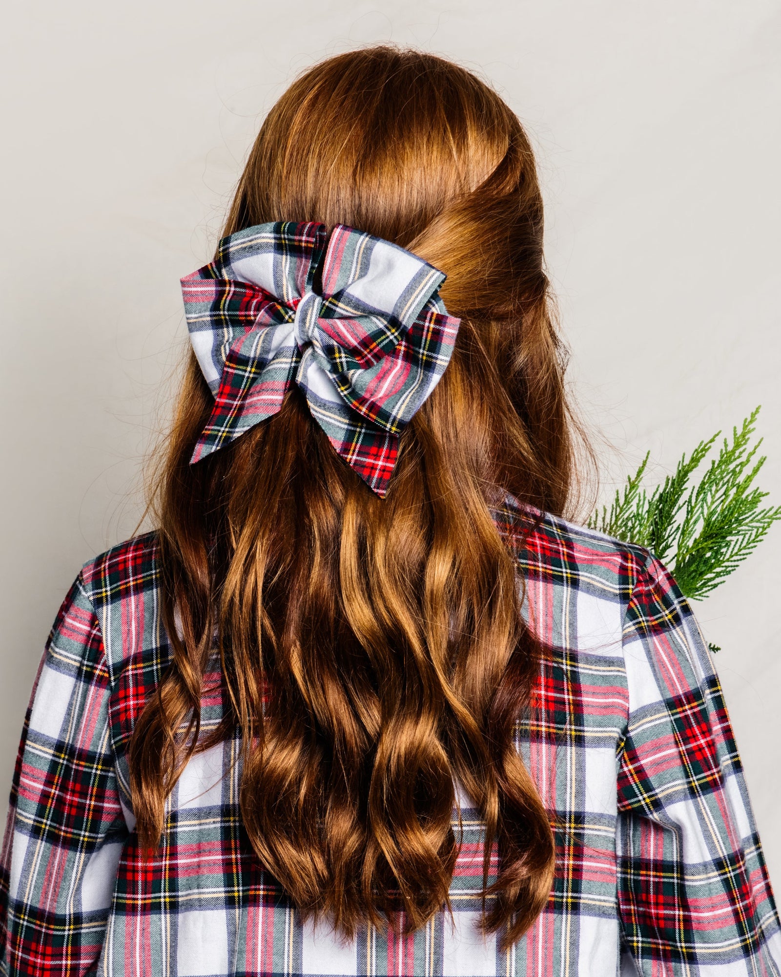 A person with long, wavy red hair, seen from behind in a plaid shirt, wears a large Petite Plume Balmoral Tartan hair bow. They hold a small bunch of green foliage, capturing the essence of girls holiday gifts.