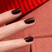 A close-up of a hand with neatly manicured nails painted in Manucurist Hollyhock, a rich black red gel polish, resting on textured red and pink fabric.