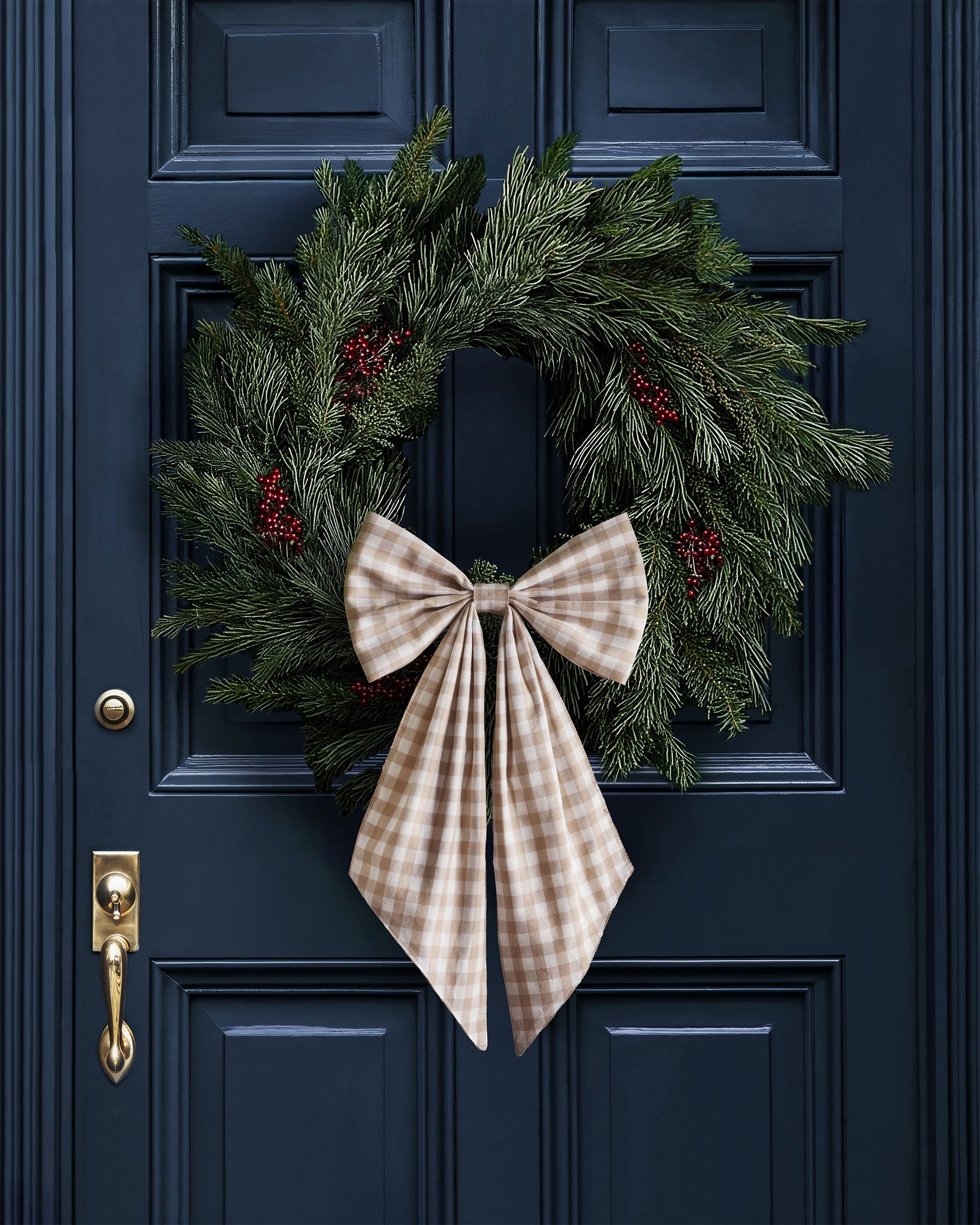 A green pine wreath with red berries and a large camel gingham bow hangs on a dark blue door with a brass handle and lock.