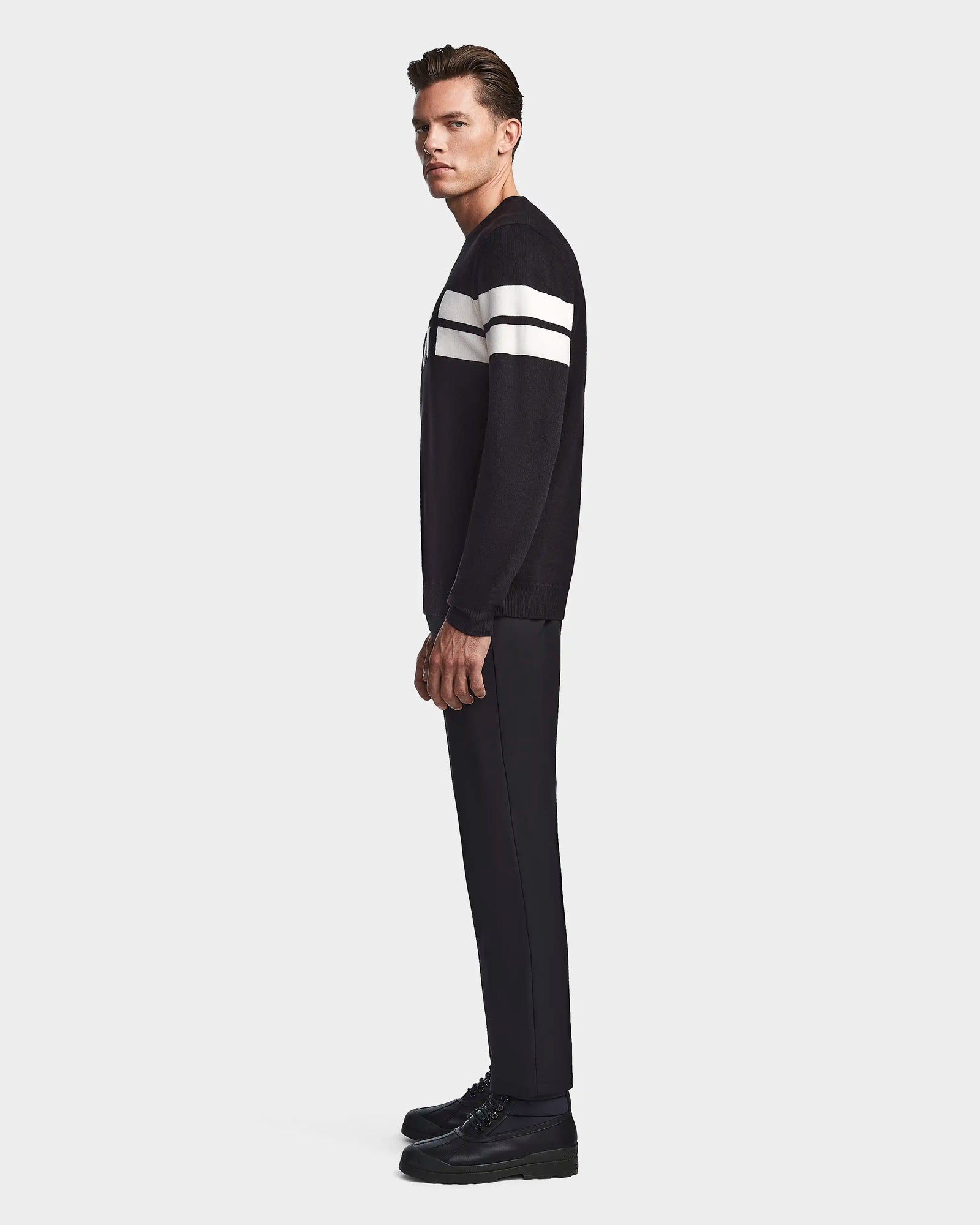 A man in profile wears the Rudsak OMAR - 6523507 Black crewneck sweater with two white sleeve stripes, paired with black pants and shoes, standing against a plain white background.