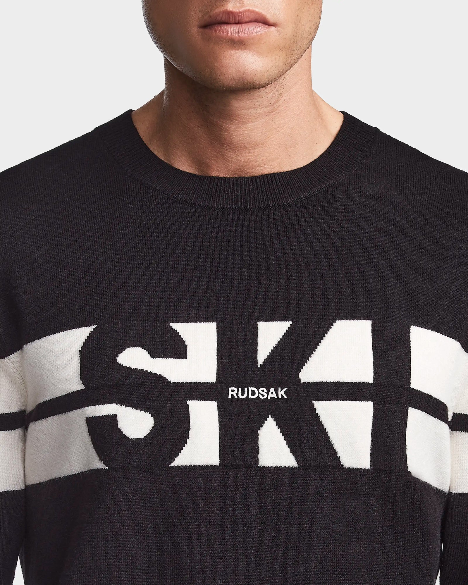 A person wears the OMAR - 6523507 Black men’s knit crewneck sweater by Rudsak, featuring bold white SK letters and the brand name across the chest. The image shows only the lower face, neck, and upper chest.