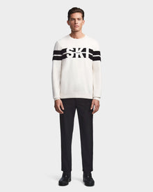 A man stands facing forward in the Rudsak OMAR - 6523507 Iceland men’s crewneck knit sweater with black horizontal stripes and SKI on the chest, paired with black pants and shoes, against a plain light background.