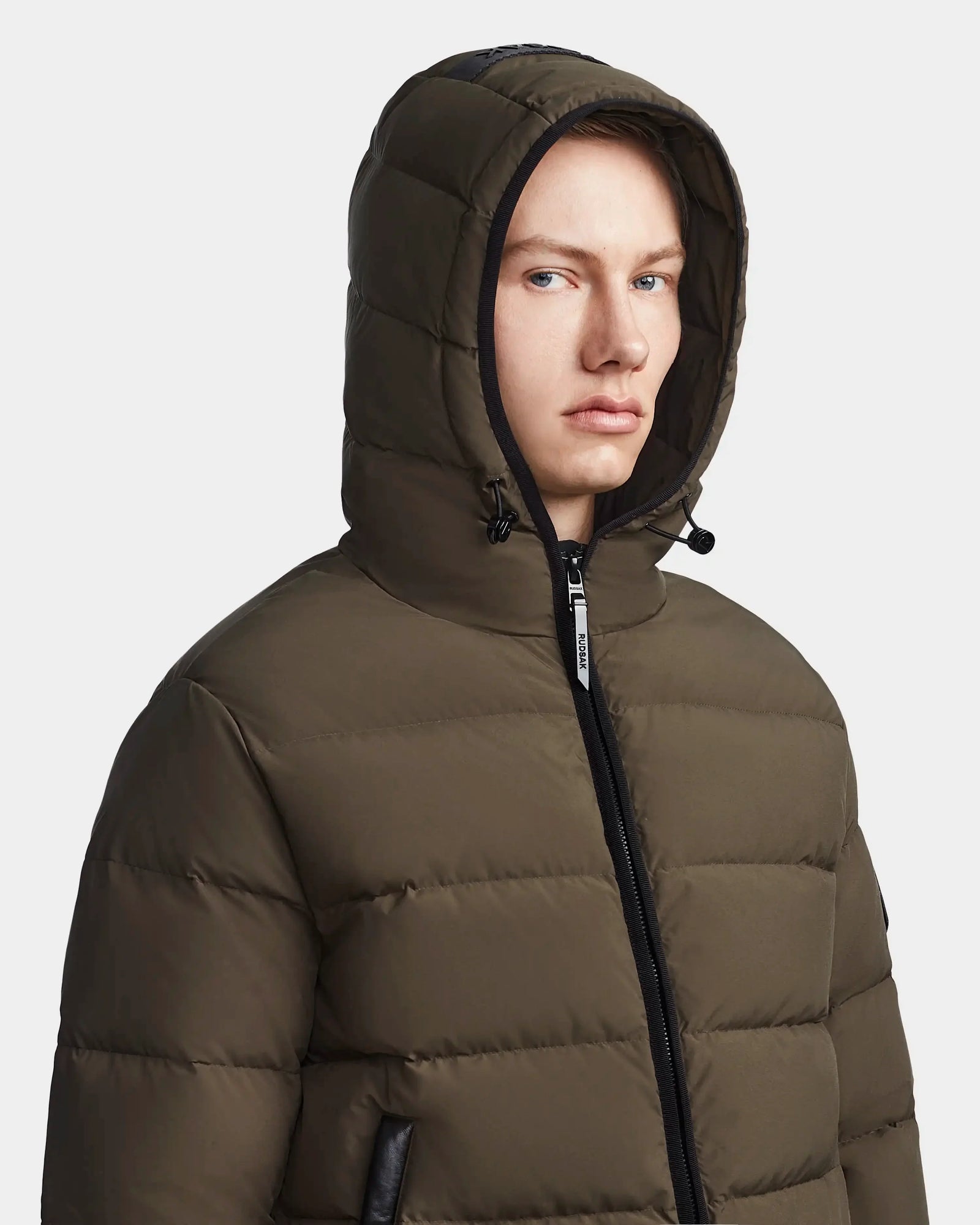 A man wears the Rudsak DEMIAN M - M123507 down puffer in light olive, featuring a hood and quilted sustainable recycled fabric, as he looks to the side against a plain light background.