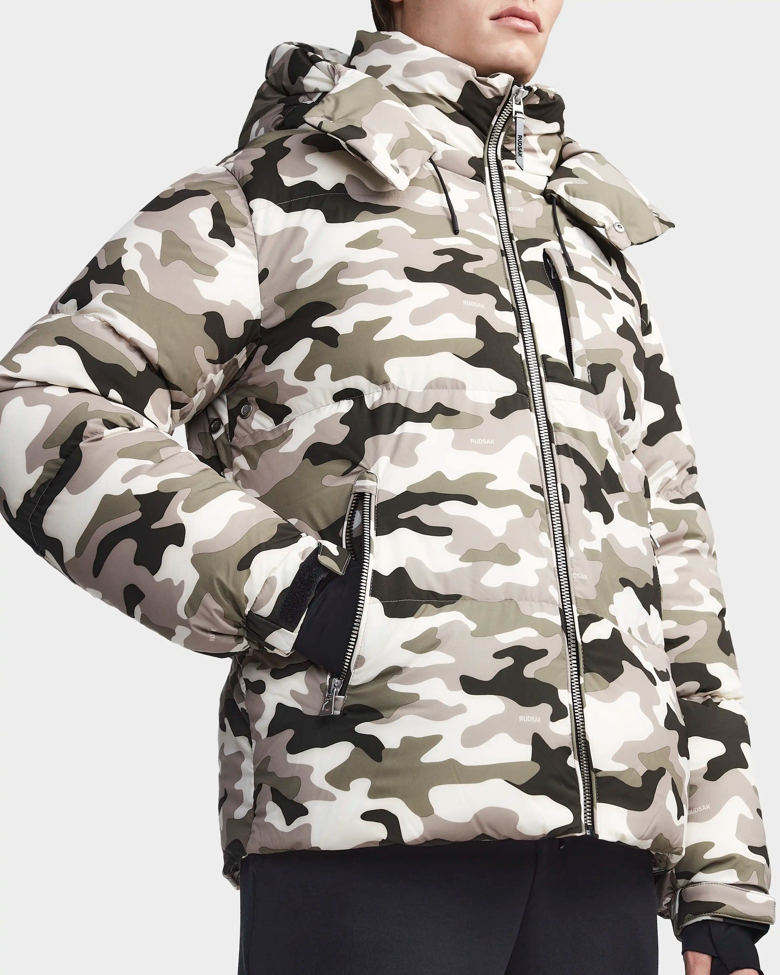 A person stands with one hand in the pocket, wearing a Rudsak VINCENT M - 6123510 Camouflage men’s down puffer jacket featuring a light beige, gray, and black pattern, lightweight insulation, and a zippered front. The face is partially out of frame.