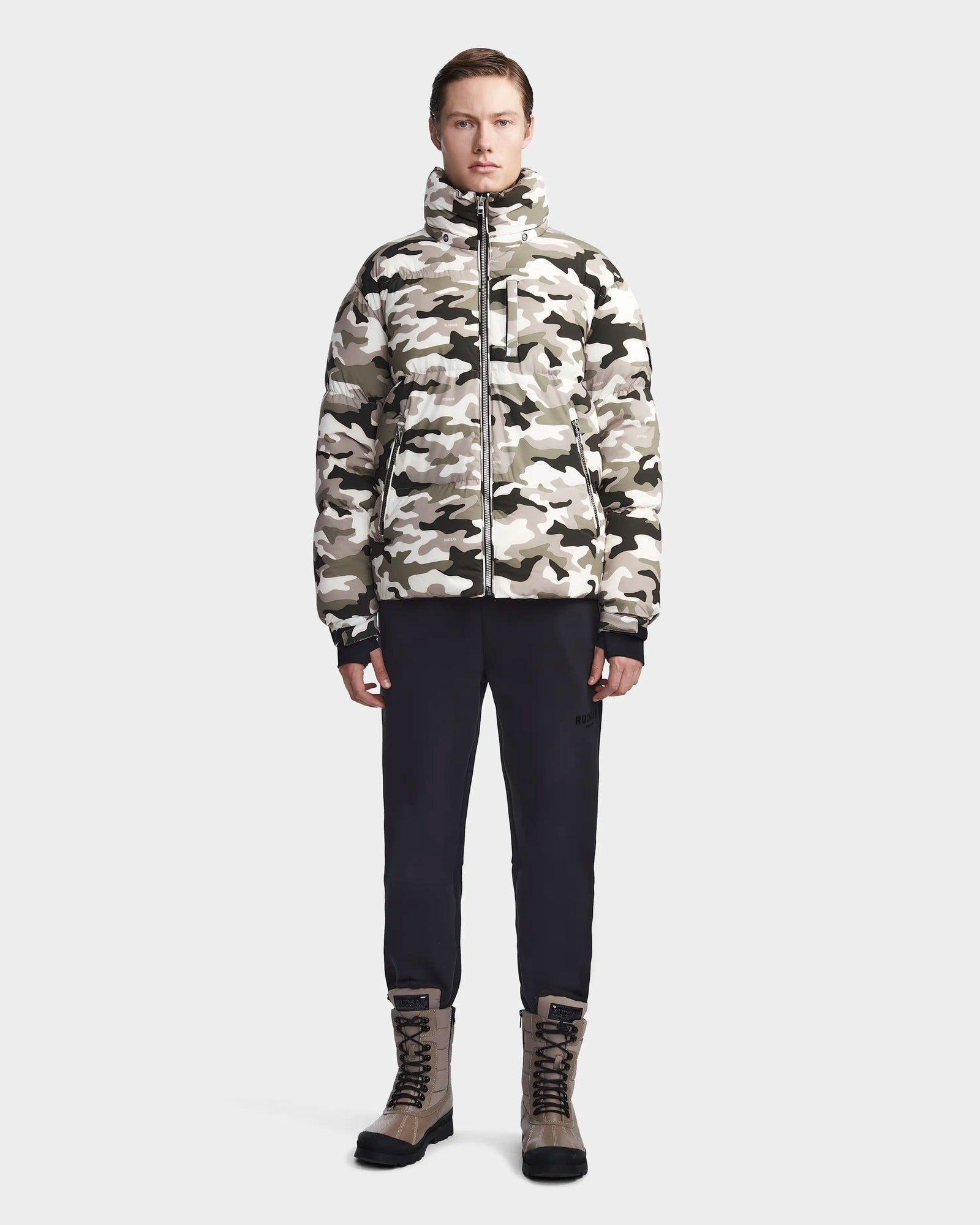 A person stands facing forward, wearing the Rudsak VINCENT M - 6123510 Camouflage men's down puffer in beige and gray camo, paired with black pants and tan lace-up boots, against a plain light gray background.