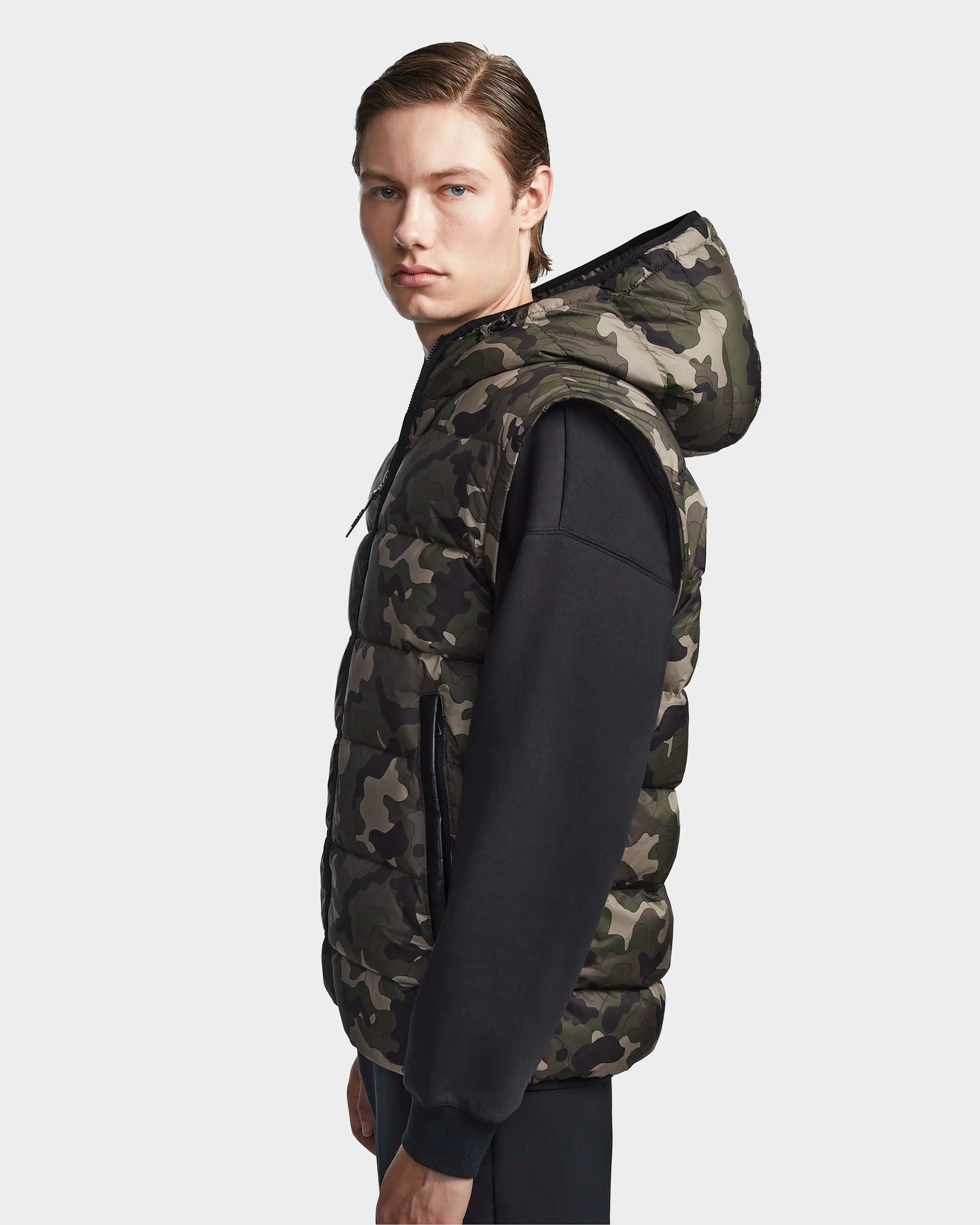 A young man with straight brown hair wears the Rudsak DEMIAN M-R - M123504 Camouflage men’s down puffer vest over a black long-sleeve shirt, standing against a plain light background and looking towards the camera.
