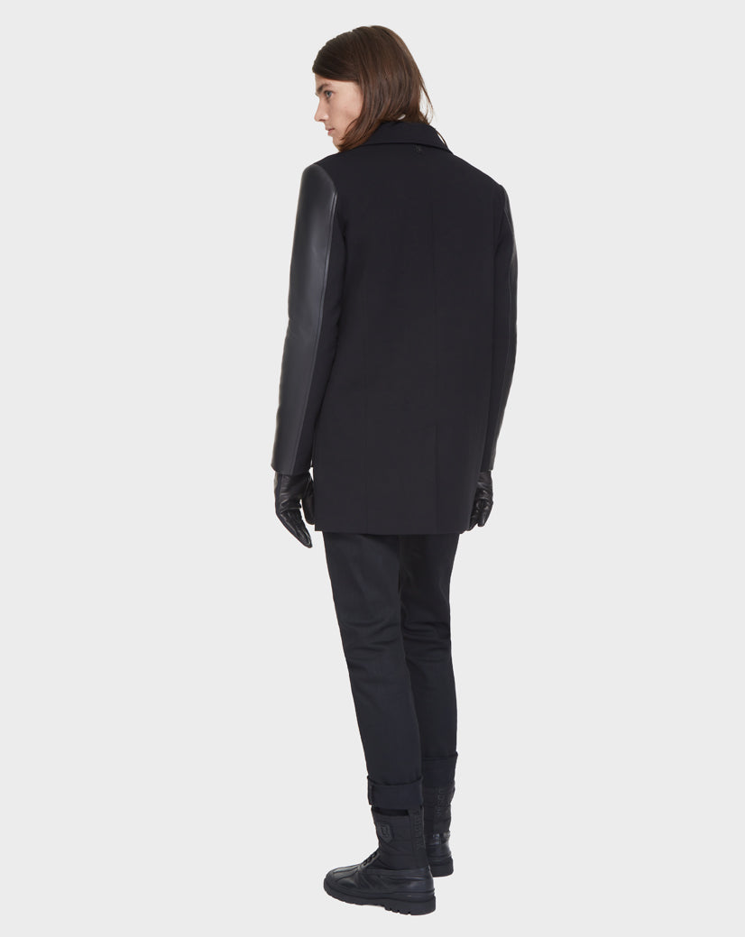 Wearing the Rudsak BEAU - M121504 BLACK coat, a person with straight, shoulder-length brown hair stands facing away from the camera against a plain light background, styled with black gloves, pants, and boots.