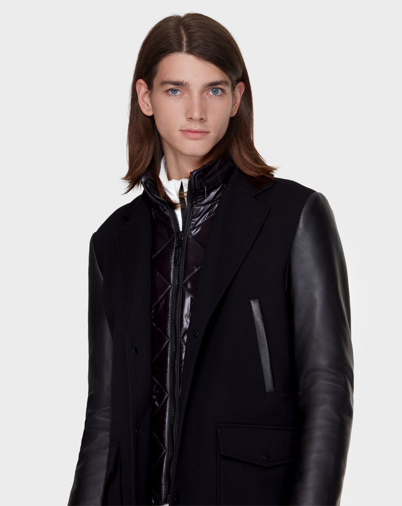 A young person with straight brown hair models the Rudsak BEAU - M121504 BLACK men’s coat with leather sleeves over a glossy black zip-up jacket, standing against a plain light background.