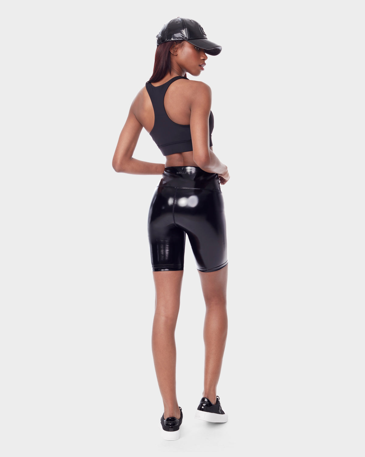 A woman faces away, wearing the BRIA - 8523016 BLACK by Rudsak—a black racerback crop top and high waist vegan leather biker shorts—with black sneakers and a cap. She looks to the side, hand on hip, against a white background.