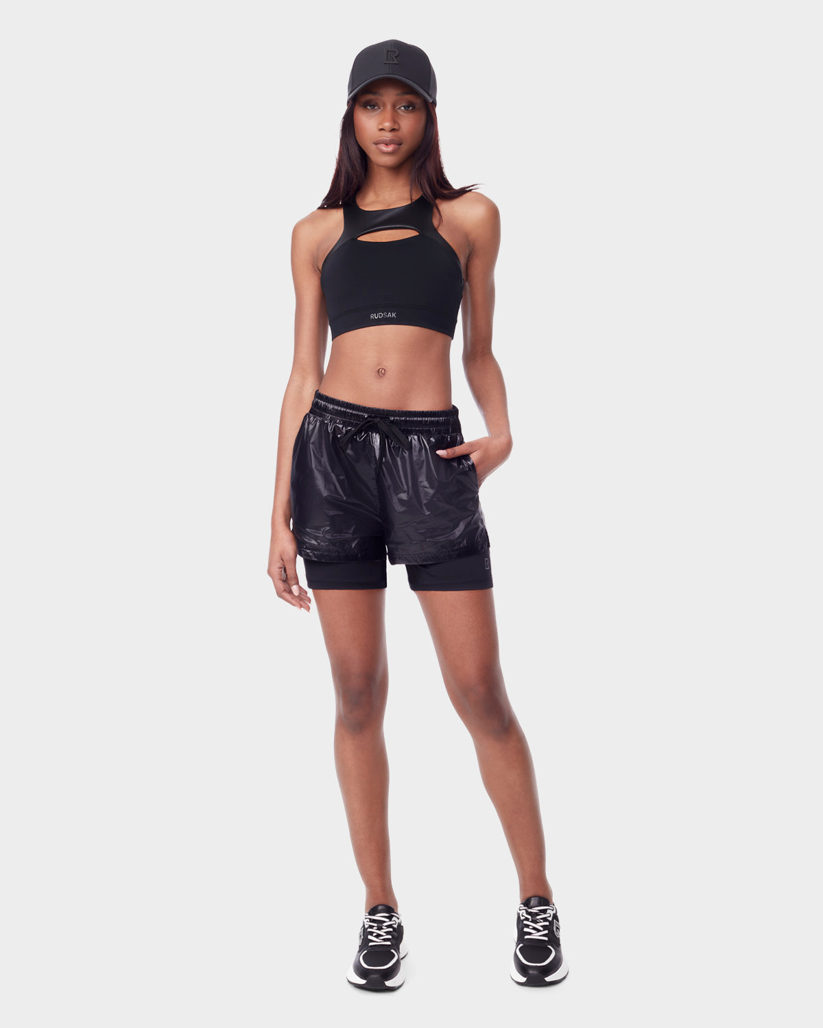 A woman wears the RYLEE - 8523001 BLACK running shorts by Rudsak, featuring recycled semi-gloss fabric and a spandex inner short, paired with a black crop top, black sneakers, and a black baseball cap against a plain light background.