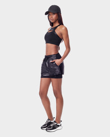 A woman stands sideways against a white background, wearing Rudsaks RYLEE - 8523001 BLACK: a black cap, sleeveless crop top, and shiny black workout shorts with pockets made from recycled fabric, paired with black-and-white sneakers.