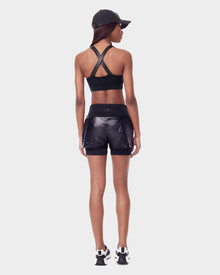 A woman stands facing away, wearing Rudsaks RYLEE - 8523001 BLACK womens running shorts made from recycled semi-gloss fabric with a spandex inner short, paired with a crisscross sports bra, black sneakers, and a cap against a plain white background.