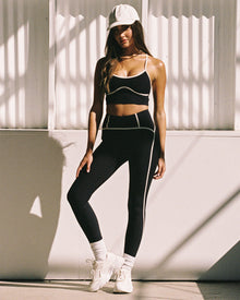 Outdoor boxing-themed editorial shot of model wearing the Revel Legging in black-cream, a high-waist legging with cream contrast piping, with matching black-cream Revel Top, white cap and chunky white sneakers.