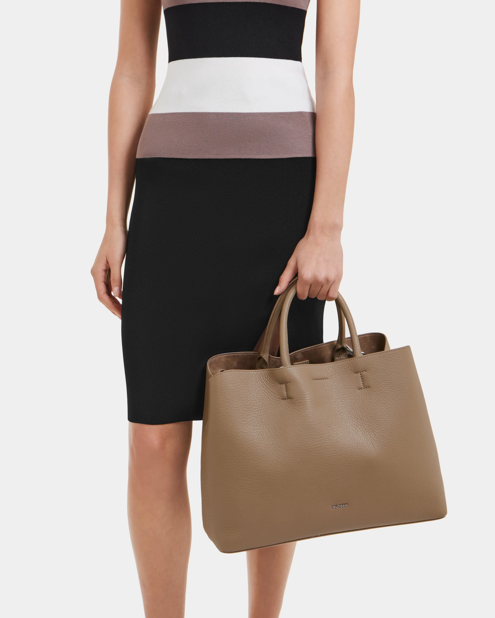A person in a sleeveless striped top and black skirt holds the Rudsak CATIRA L - 8325007 Almond women’s tote, showcasing its two handles as a statement accessory against a plain white background.