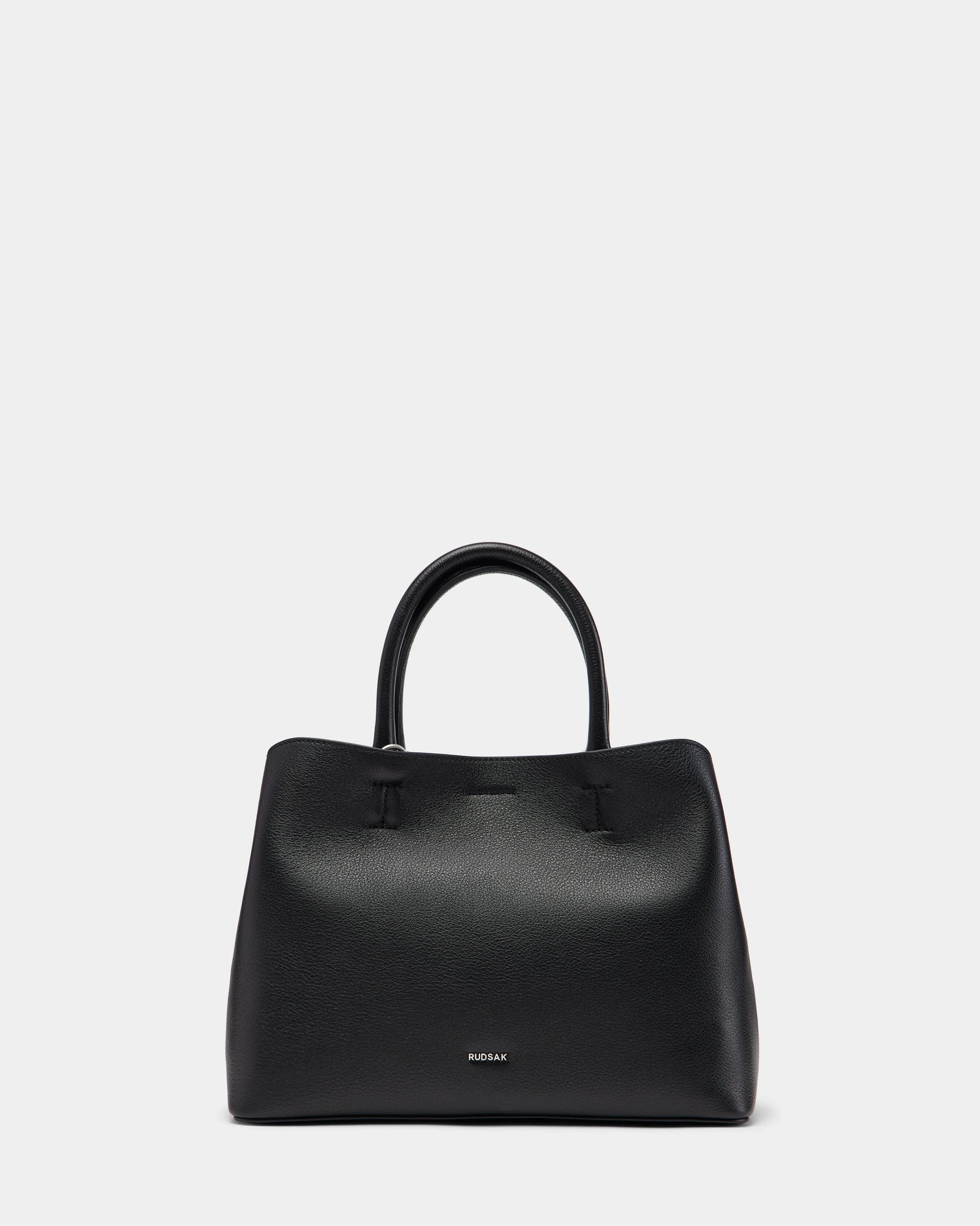 The CATIRA S - 8325006 Black by Rudsak is a structured black pebble leather tote with two handles, minimal seams, and a removable shoulder strap. The Rudsak logo appears in small silver lettering near the bottom center on a plain white background.
