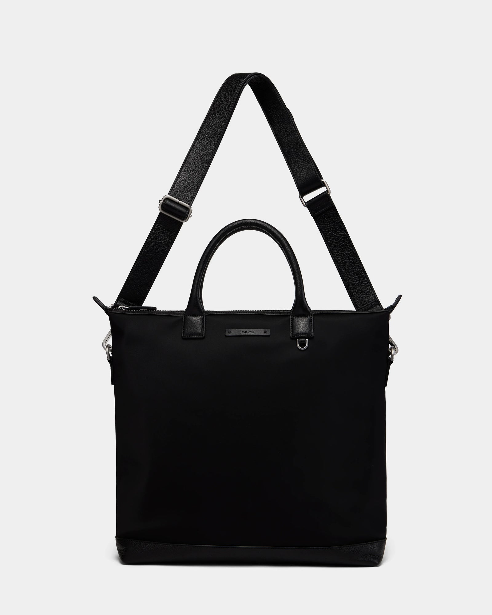 The DEXTER - 9324513 Black by Rudsak is a versatile unisex canvas tote with short handles, an adjustable shoulder strap, a rectangular shape, and a small metal nameplate on the front, shown against a plain white background.