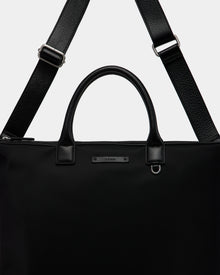 A close-up of the Rudsak DEXTER - 9324513 Black tote, a sleek unisex bag with sturdy leather handles, a detachable shoulder strap, and a small metallic brand label on the front.