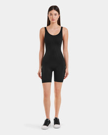 A woman with straight brown hair wears the Rudsak KAILEY - 8523012 Black Pattern fitted sleeveless bodysuit with shorts and black sneakers, standing against a plain white background, facing forward with arms relaxed at her sides.