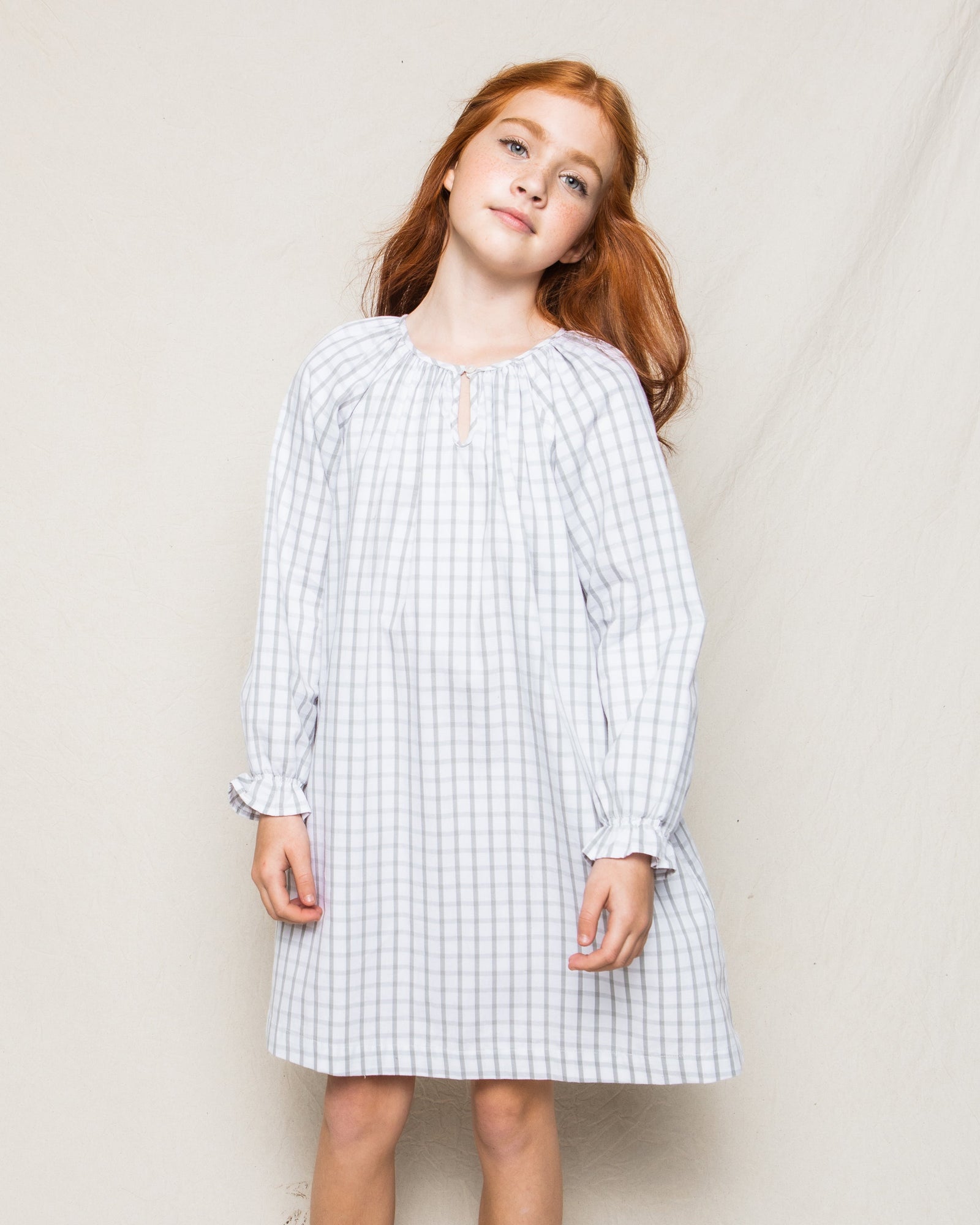A young girl with long red hair stands against a plain backdrop in Petite Plumes Girls Twill Delphine Nightgown in Regent Tattersall, reminiscent of luxurious sleepwear. She tilts her head slightly, gazing forward with serene confidence.