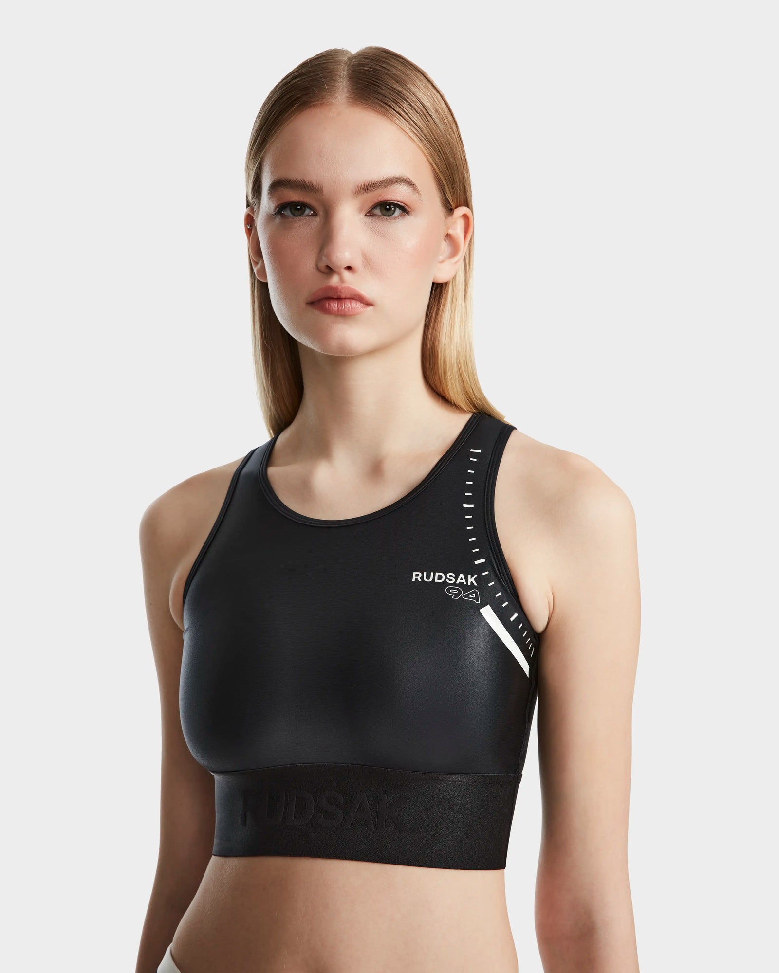 A young woman with straight, blonde hair wears the Rudsak TRACY - 8524016 Black high-performance racer-back sports bra with white logo details, standing against a plain white background and looking at the camera.