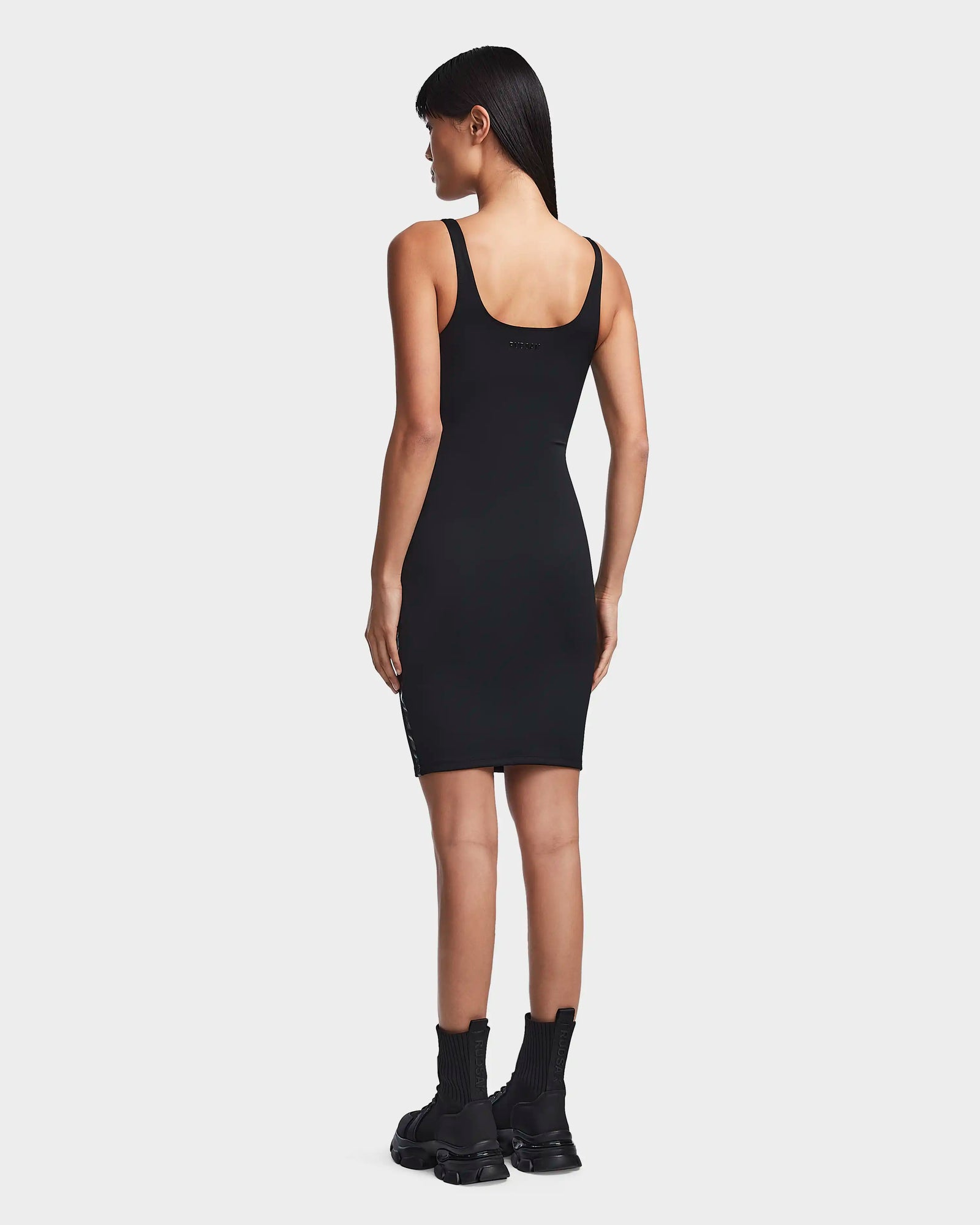 A woman with straight dark hair faces away, wearing the Rudsak MILEY - 8523034 BLACK bodycon dress and black chunky boots with high socks, standing against a plain white background.