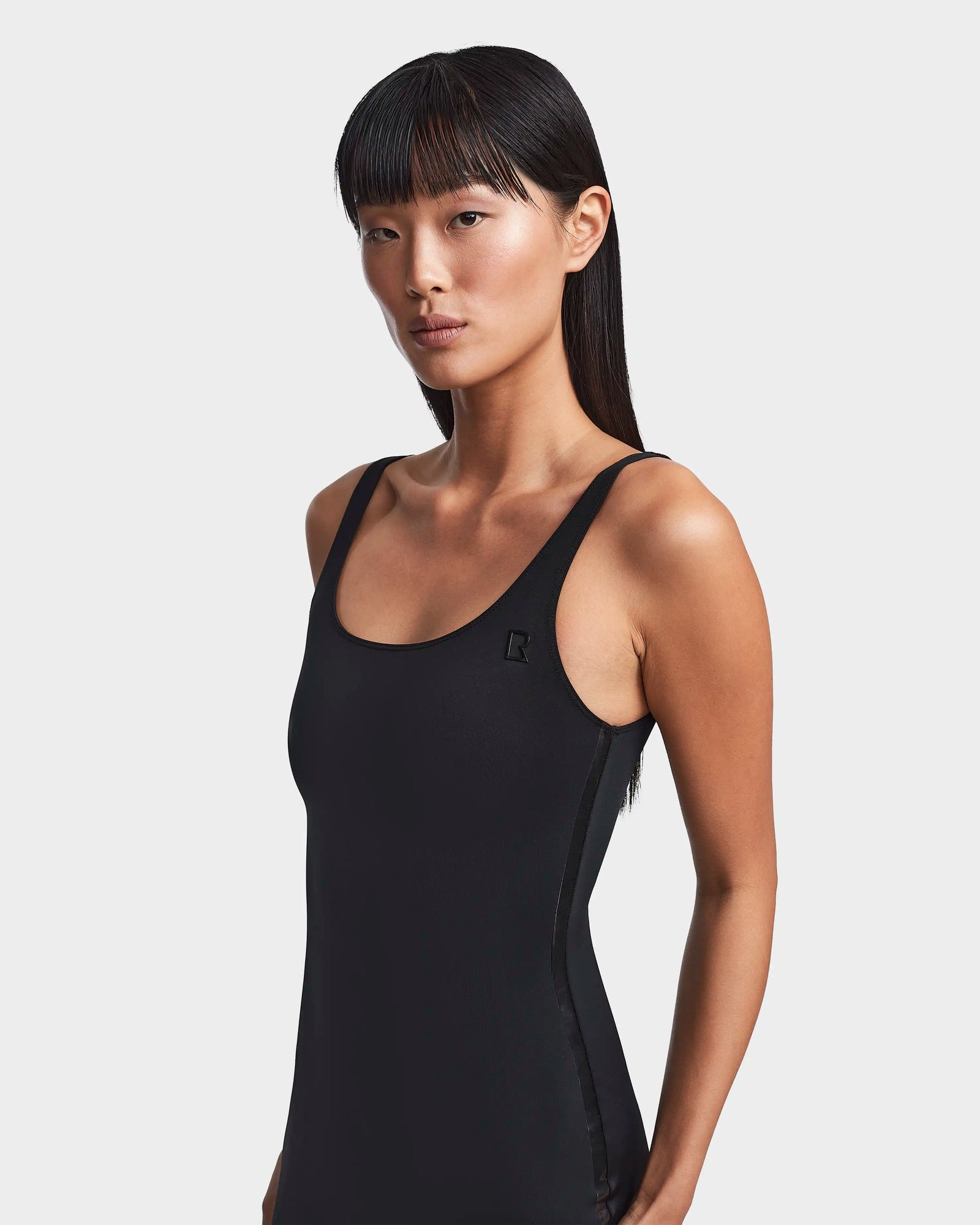 A woman with long, straight black hair and bangs confidently models the Rudsak MILEY - 8523034 BLACK fitted tank top against a plain white background.