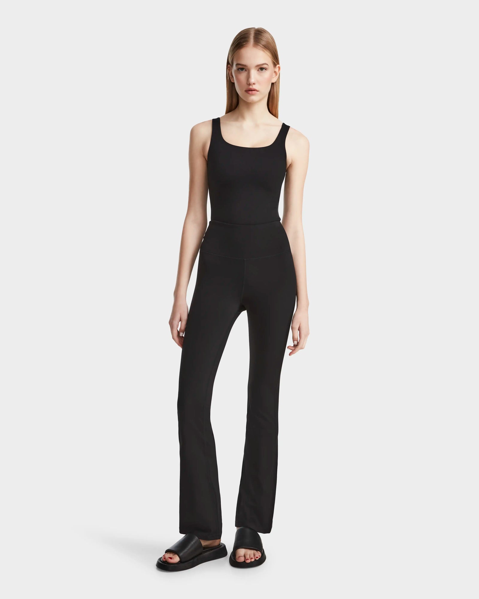 A woman with straight blonde hair and a neutral expression wears Rudsaks ADRIANA - 8524009 Black fitted technical flared-leg leggings, a black tank top, and black slide sandals against a plain white background.