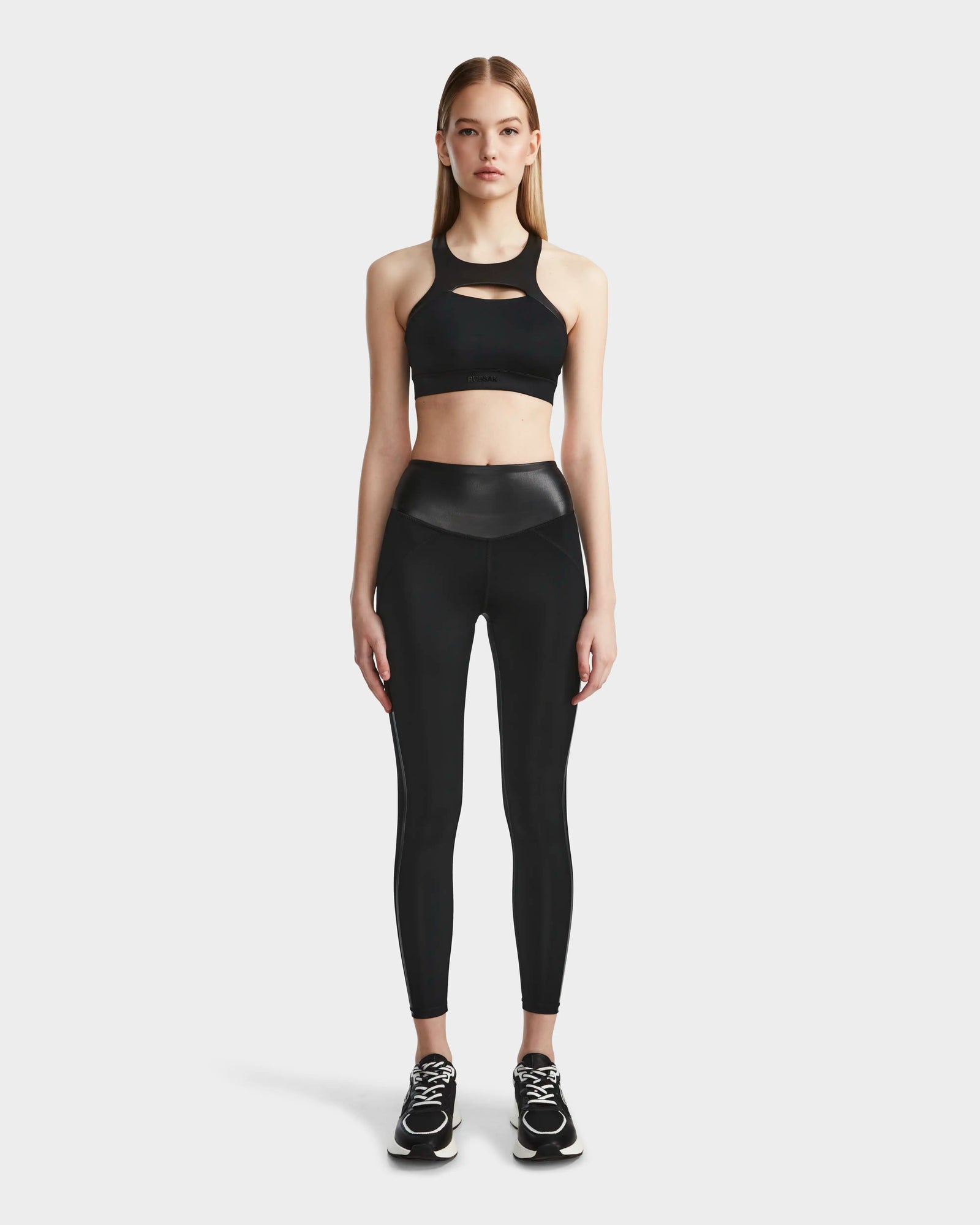 A woman faces forward wearing Rudsak’s THEA - 8523003 BLACK high-rise leggings with a vegan leather waistband, a matching sports bra, and black athletic shoes against a plain white background.