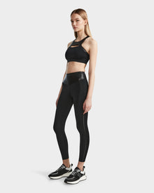 A woman with straight light brown hair and a neutral expression stands against a plain background wearing THEA - 8523003 BLACK high-rise womens leggings by Rudsak, featuring a vegan leather waistband, paired with a sporty crop top and black sneakers.
