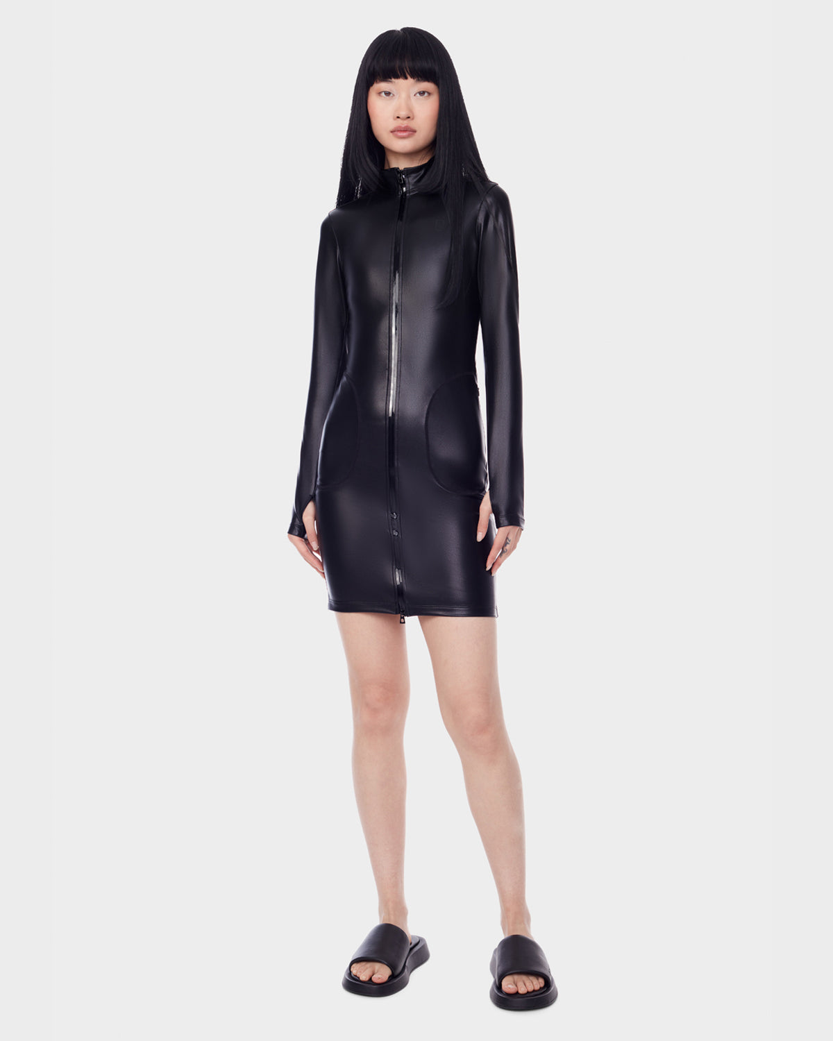 A woman with straight black hair and bangs wears the Rudsak SHANA - 8522508 BLACK long-sleeve stretch vegan leather zip-up mini dress with black slide sandals, standing against a plain white background.