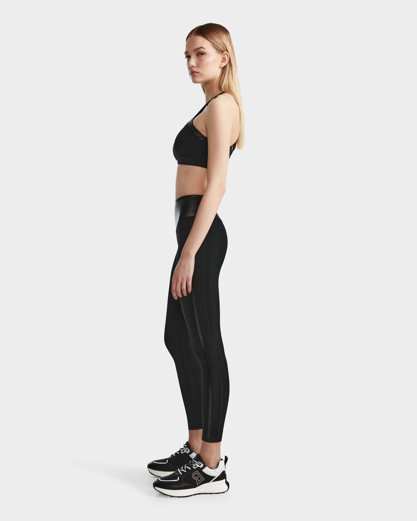 A woman stands in profile wearing the RUDSAK ARYA - 8523002 BLACK sports bra, paired with high-waisted black leggings and black sneakers with white soles, set against a plain white background.