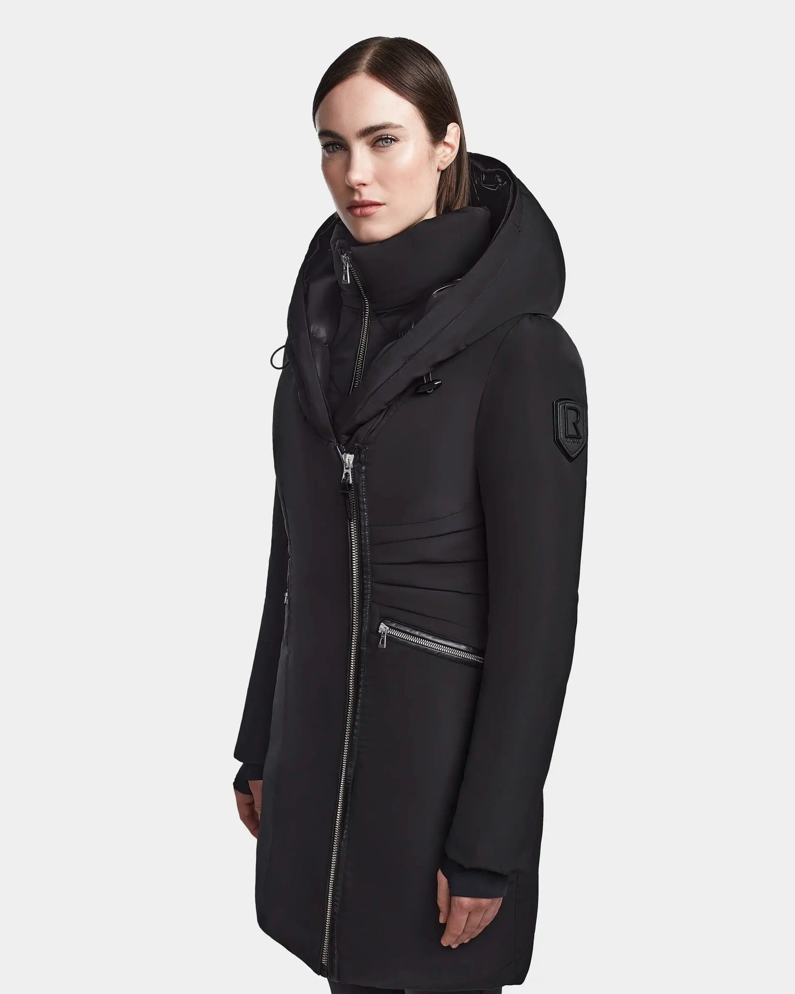 A woman with straight dark hair wears the Rudsak CORA - L123538 Black women's fitted parka, a long black sustainable winter coat with a high collar, large hood, and silver zippers, standing against a plain light gray background.
