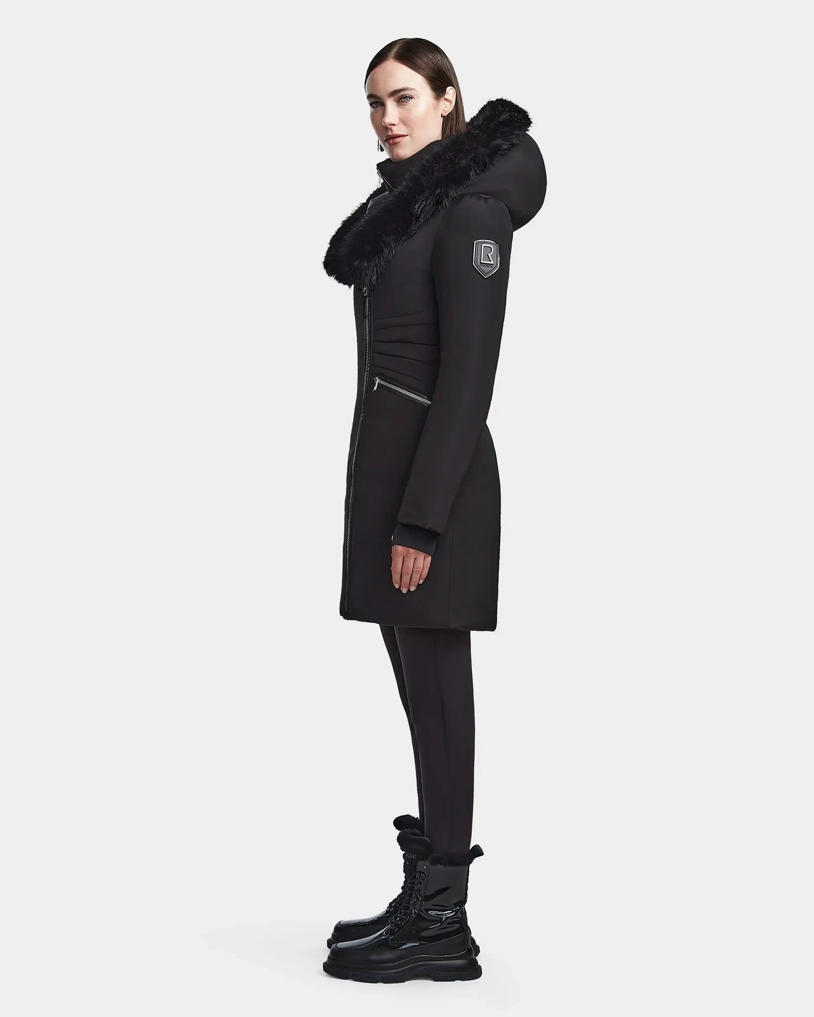 A woman stands in profile against a plain light background, wearing the Rudsak CORA - L123538 Black women’s fitted parka with a fur-trimmed hood, paired with black leggings and black lace-up boots.