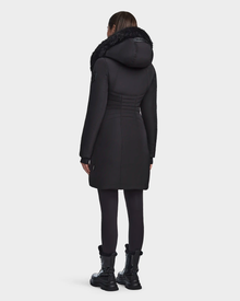 A person wearing the Rudsak CORA - L123538 Black women’s fitted parka—a long black sustainable winter coat with a fur-lined hood—plus black leggings and boots is shown from the back against a plain white background.