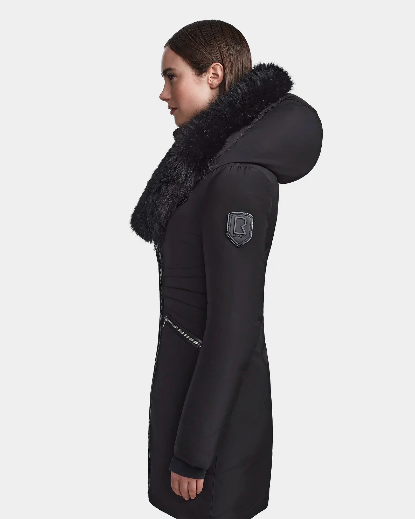 A woman in profile wears the Rudsak CORA - L123538 Black, a long sustainable women’s parka with a large black fur-trimmed hood, visible arm badge, and side pocket zipper. The background is plain white.