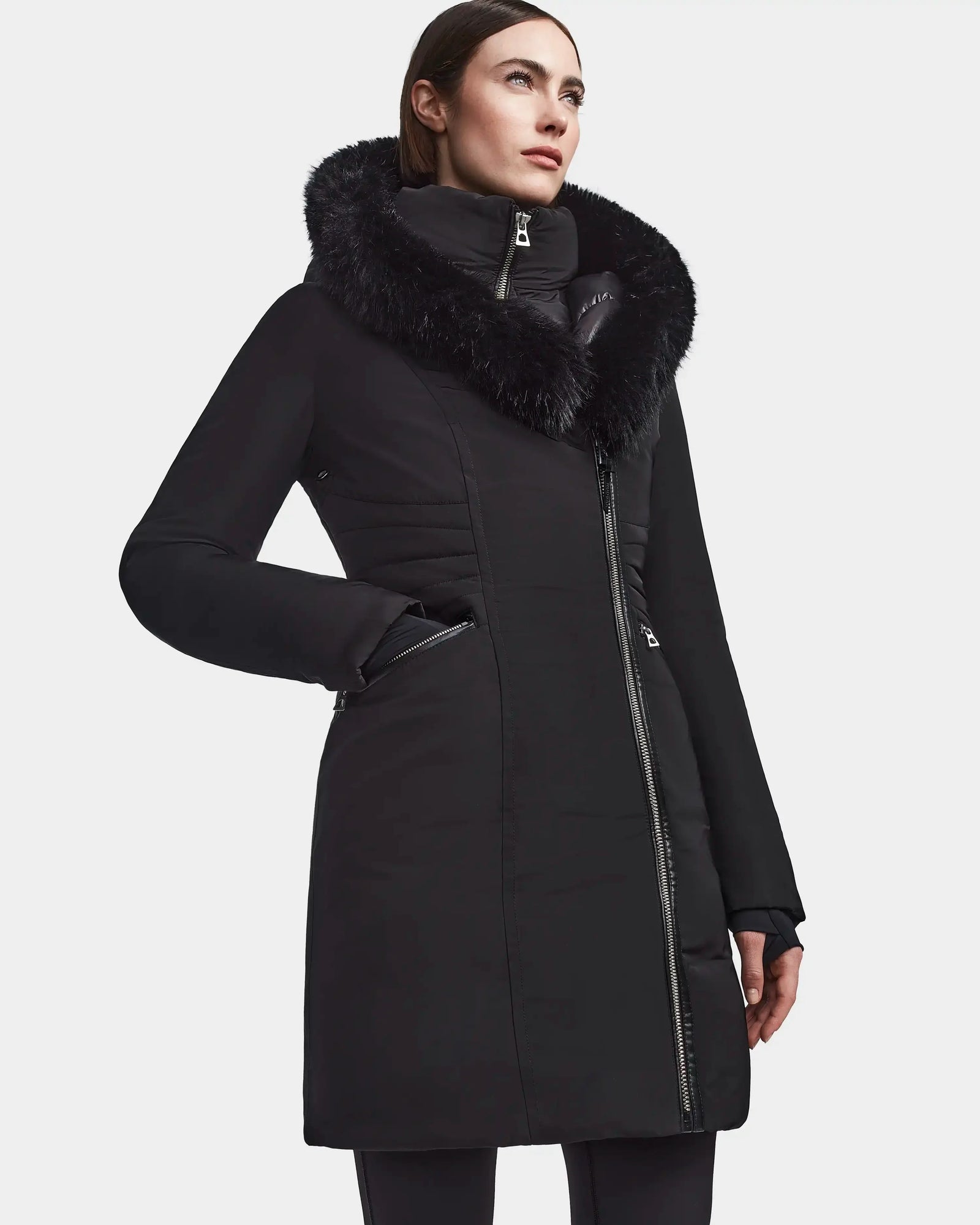A person wears the Rudsak CORA - L123538 Black women’s fitted parka, a long black winter coat with a fluffy faux fur collar and asymmetrical zip closure, standing against a plain white background.