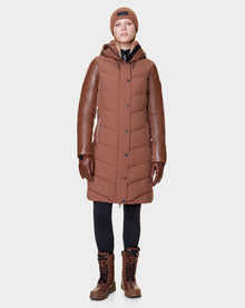 A person wears the Rudsak ELSA - L121519 TAUPE women’s quilted down puffer coat with leather sleeves, plus brown gloves, boots, a knit beanie, and black leggings—showcasing sustainable design—against a plain white background.