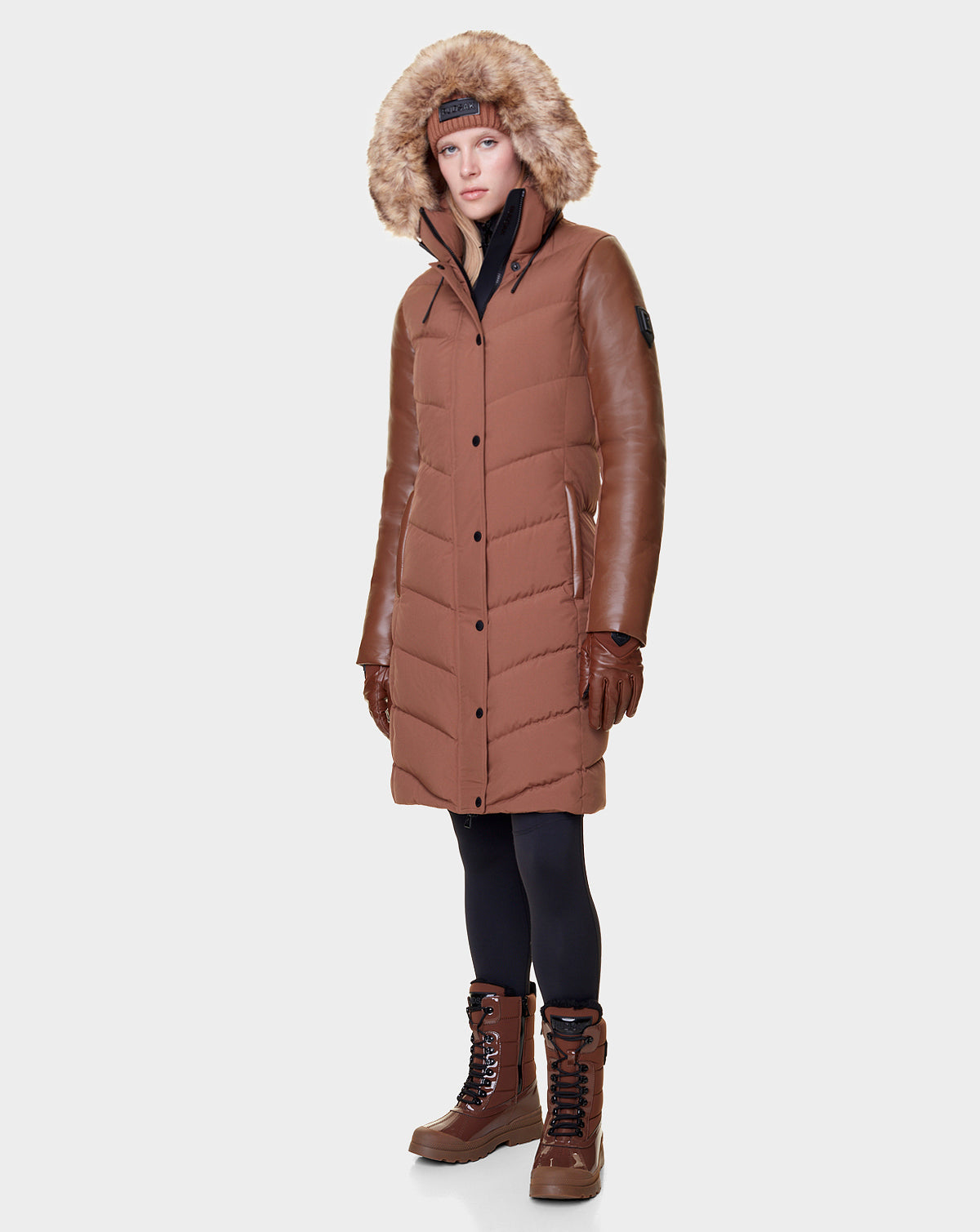 A woman faces forward wearing the Rudsak ELSA - L121519 TAUPE long brown quilted down puffer winter coat with a faux fur-lined hood, matching gloves, black leggings, and brown lace-up winter boots.