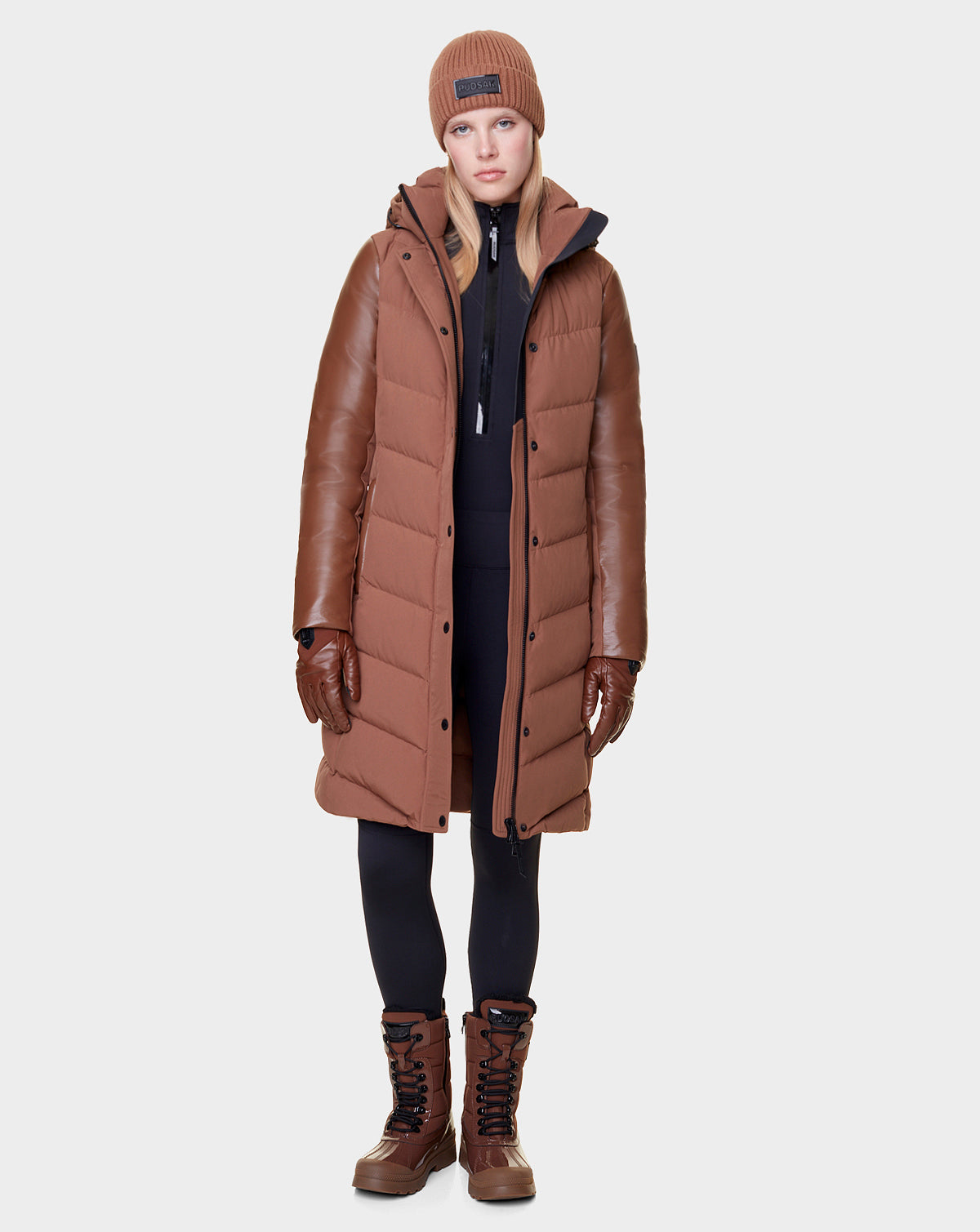A woman stands facing forward in the Rudsak ELSA - L121519 TAUPE, a long taupe women’s quilted down puffer with leather sleeves—an elegant, sustainable look paired with brown gloves, knit beanie, black leggings, and brown winter boots.