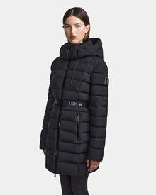 A woman models the Rudsak FRIDA M - L123514 Black quilted down jacket, featuring a belted waist, zippered front, and removable hood, against a plain white background.