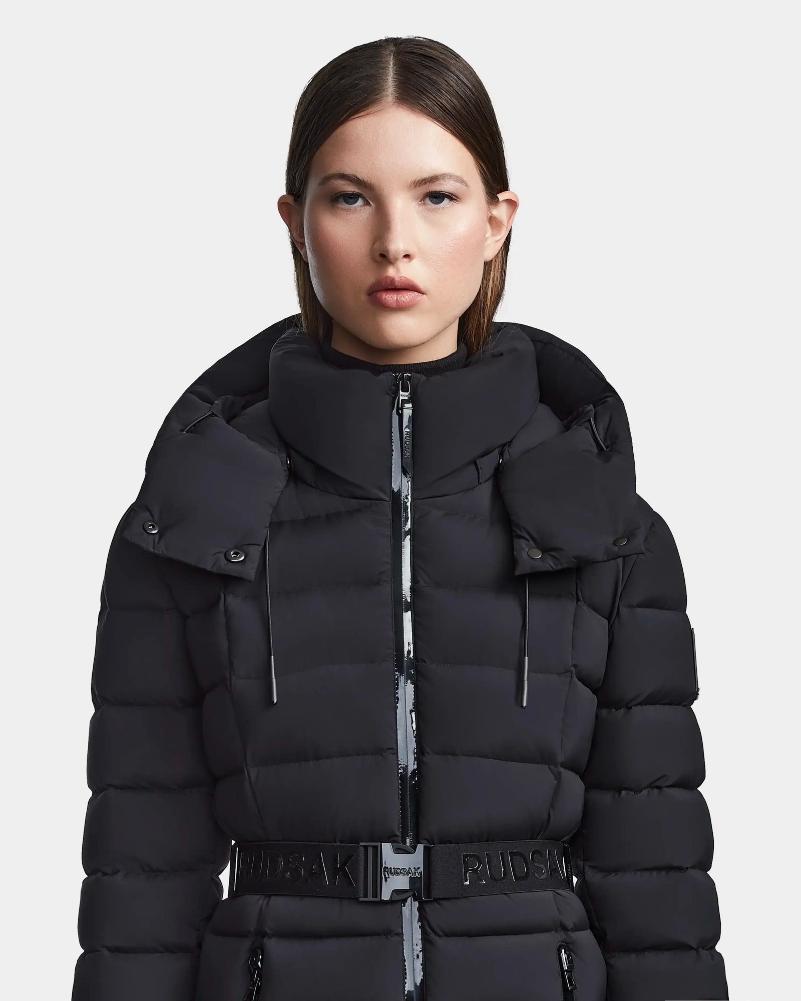 A woman with straight brown hair faces forward against a white background, wearing the Rudsak FRIDA M - L123514 Black: a black, quilted down jacket featuring a belt and removable hood.