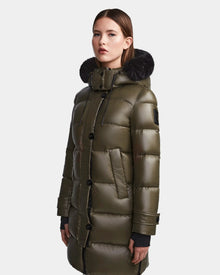 A woman stands facing forward against a plain background, wearing the Rudsak MEYA S - L123521 Light Olive down puffer with a black fur-lined hood.