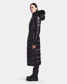 A woman stands sideways wearing the Rudsak ELSIE S - L123518 Black maxi down puffer with a fur-trimmed hood, paired with black leggings and lace-up winter boots, against a plain light background.
