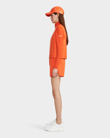 A woman stands in profile against a white background, wearing Rudsak’s LOUISE - 8124046 Orange Sorbet lightweight quilted jacket, matching shorts, an orange cap, and white platform sneakers.