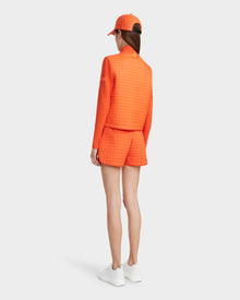 A person faces away, wearing the Rudsak LOUISE - 8124046 Orange Sorbet lightweight quilted jacket with matching shorts and a baseball cap, paired with white sneakers, against a plain white background.
