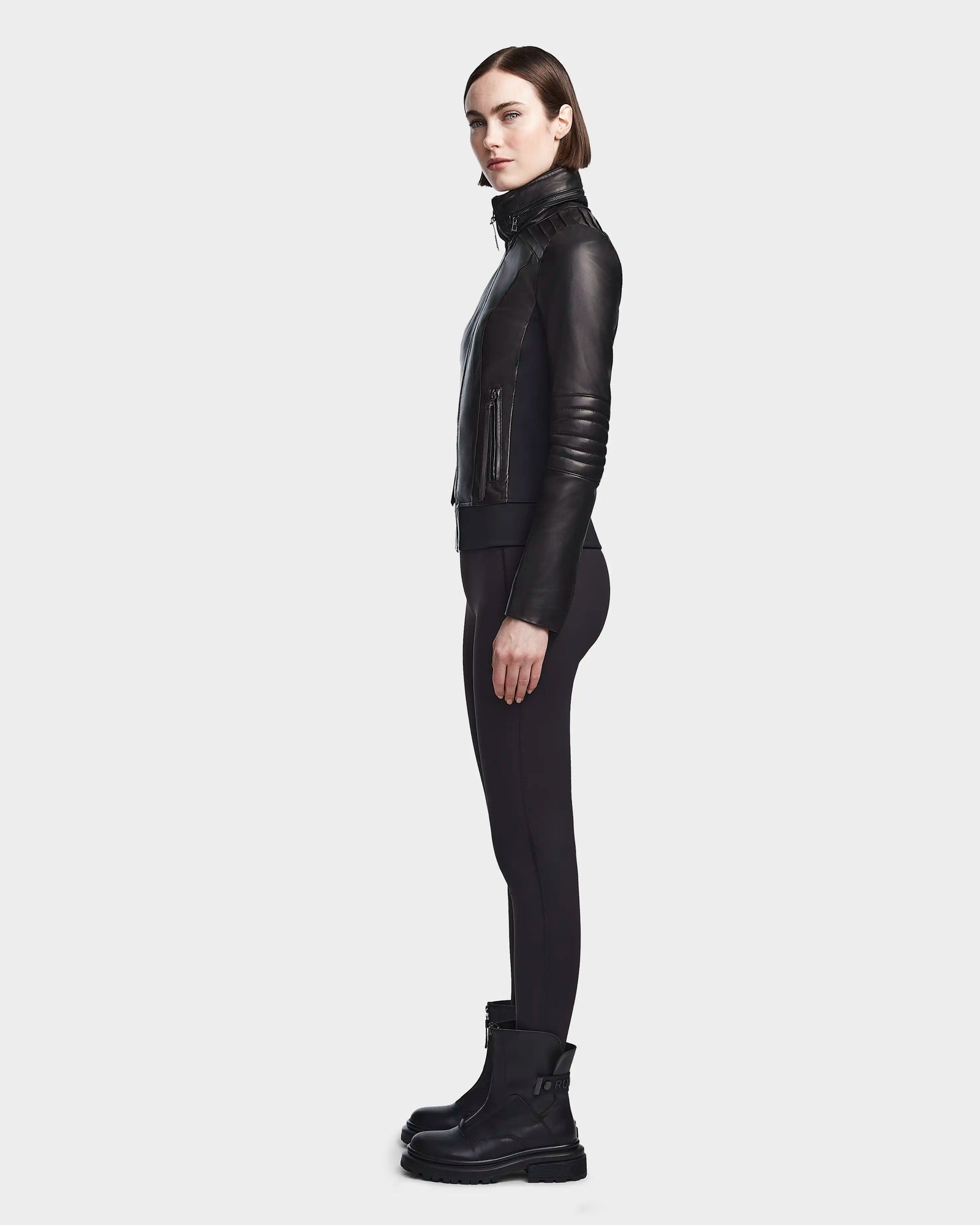 A woman stands in profile against a plain background, wearing the PRESLEY - 8122027 BLACK leather jacket by Rudsak, paired with black leggings and boots. Her straight hair is tucked behind her ears.
