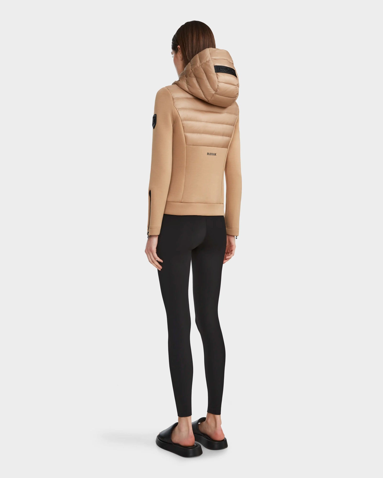 A person stands facing away, wearing the RUDSAK MABS - 8122020 TOFFEE beige lightweight jacket with waterproof zippers, black leggings, and black slip-on shoes against a plain white background.
