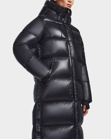 A woman stands against a white background wearing the Rudsak RITA L-S - L123526 Black maxi-length down puffer, featuring a shiny finish, high collar, hood, and large quilted horizontal sections.